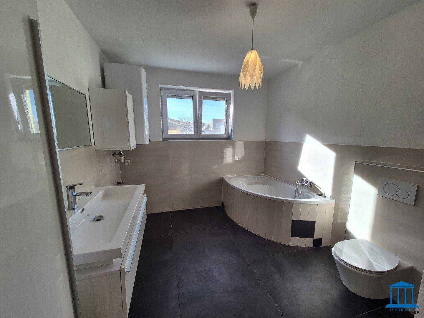Modernes Badezimmer mit dunklen Fliesen, Eckbadewanne, wandhängendem WC und Fenster für Tageslicht.