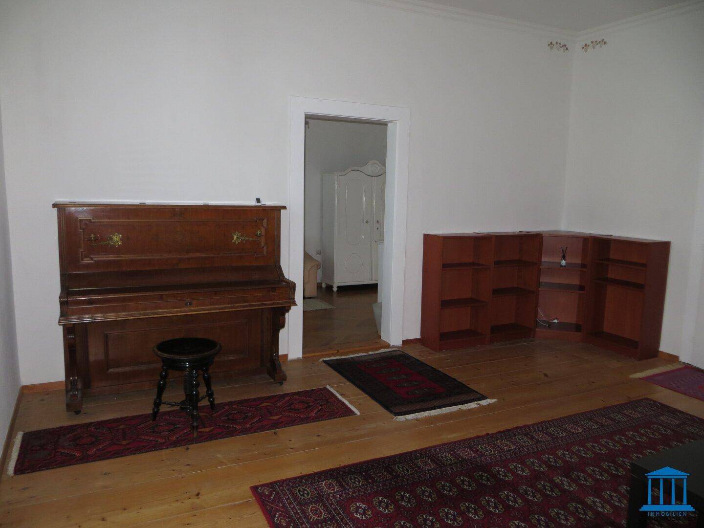 Helles Wohnzimmer mit Holzdielenboden, einem Klavier und Regalen, offen zu einem weiteren Raum.