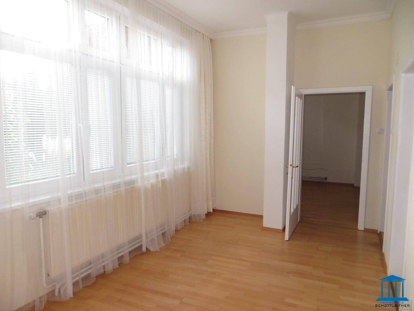 Helles Zimmer mit Parkettboden und großen Fenstern, die viel Tageslicht hereinlassen.