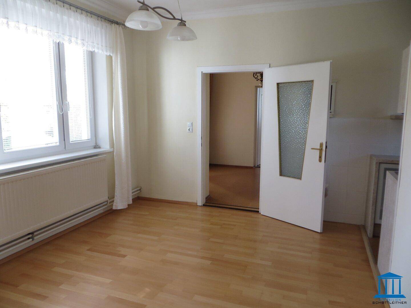 Helles Zimmer mit Parkettboden und Fenster, das zu einem weiteren Raum führt.
