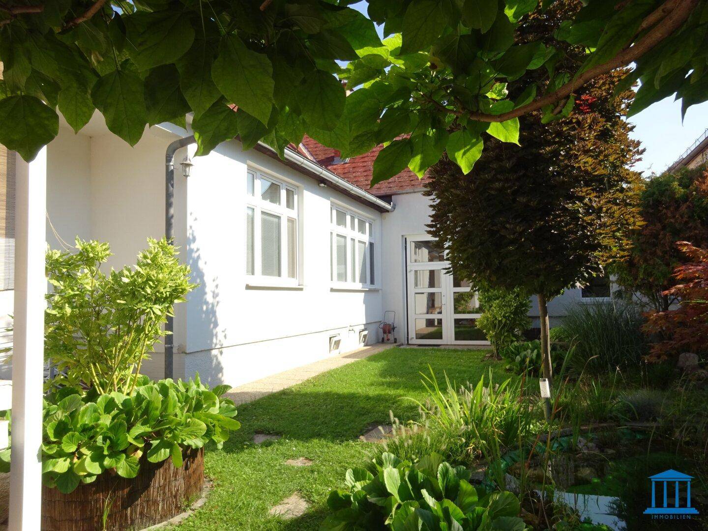 Seitenansicht des Hauses mit gepflegtem Rasen und üppiger Bepflanzung unter blauem Himmel.
