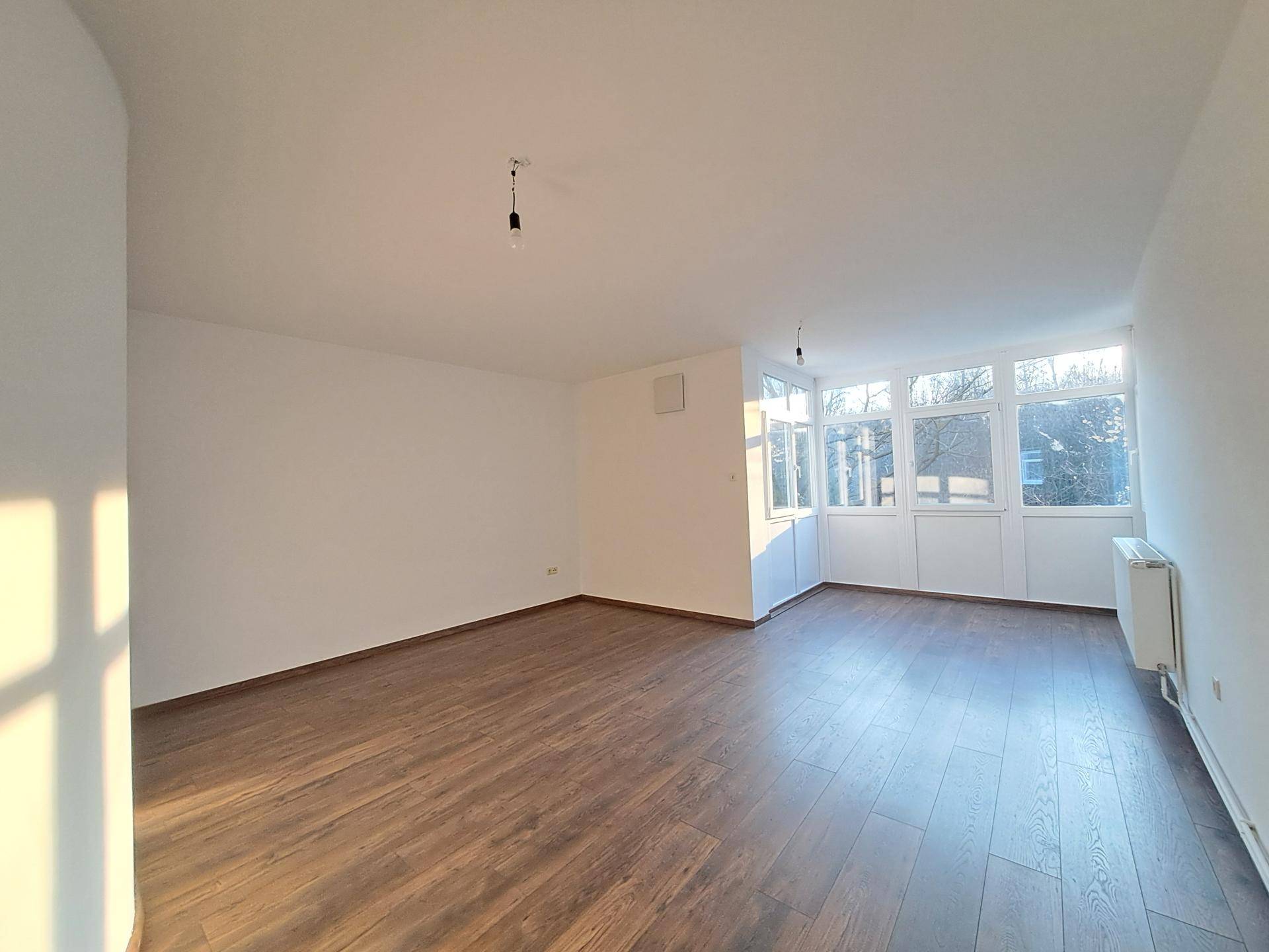 Großes, helles Wohnzimmer mit dunklem Laminatboden und einer Fensterfront, die viel Sonnenlicht hereinlässt.