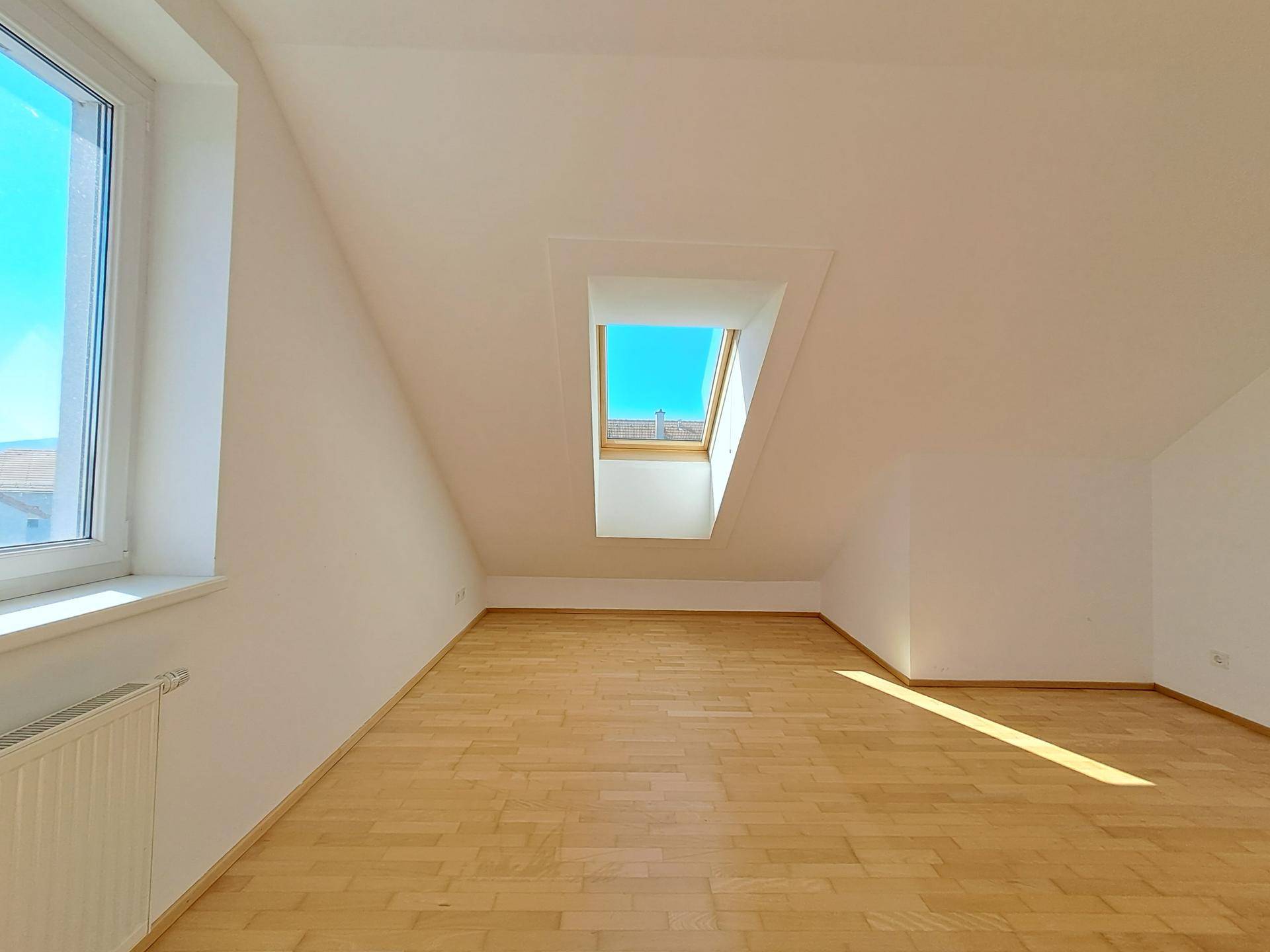Lichtdurchflutetes Zimmer im Dachgeschoss mit Holzboden und Ausblick durch Fenster und Dachfenster.