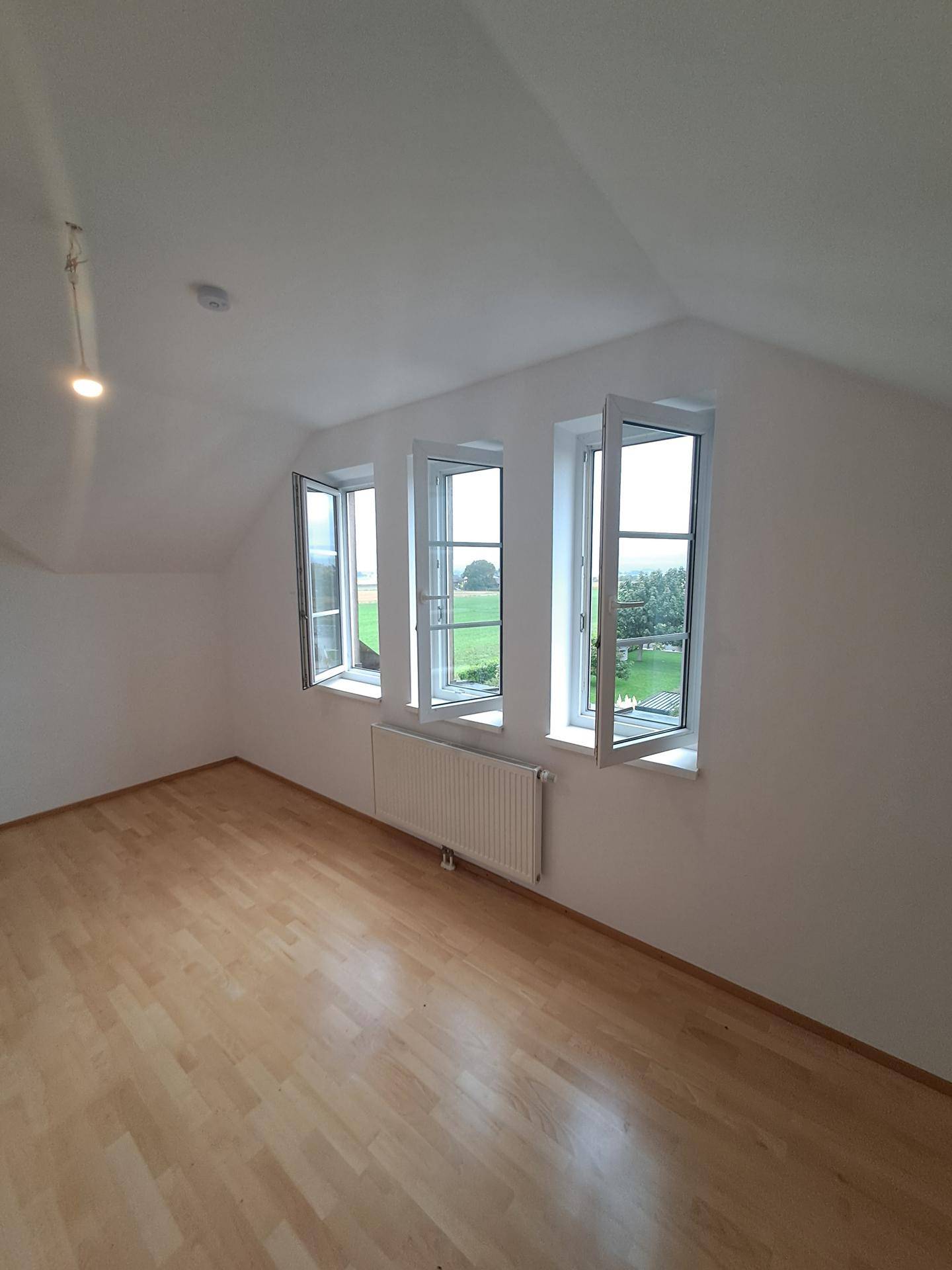 Helles Dachgeschosszimmer mit hellem Laminatboden und drei Fenstern mit Blick ins Grüne.