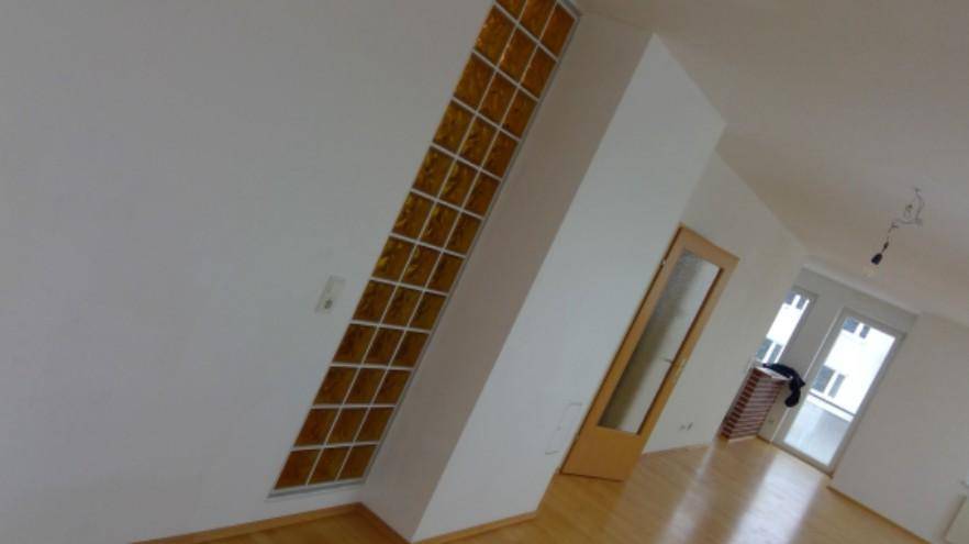 Heller Raum mit Holzfußboden, schrägen Wänden und einem dekorativen Glasbausteinfenster.