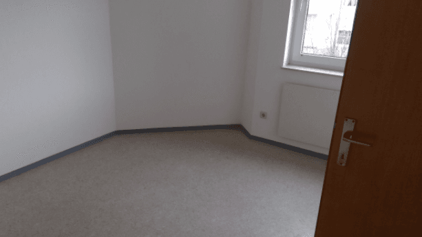 Leeres Schlafzimmer mit hellem Bodenbelag, weiß gestrichenen Wänden, Fenster und Heizkörper.