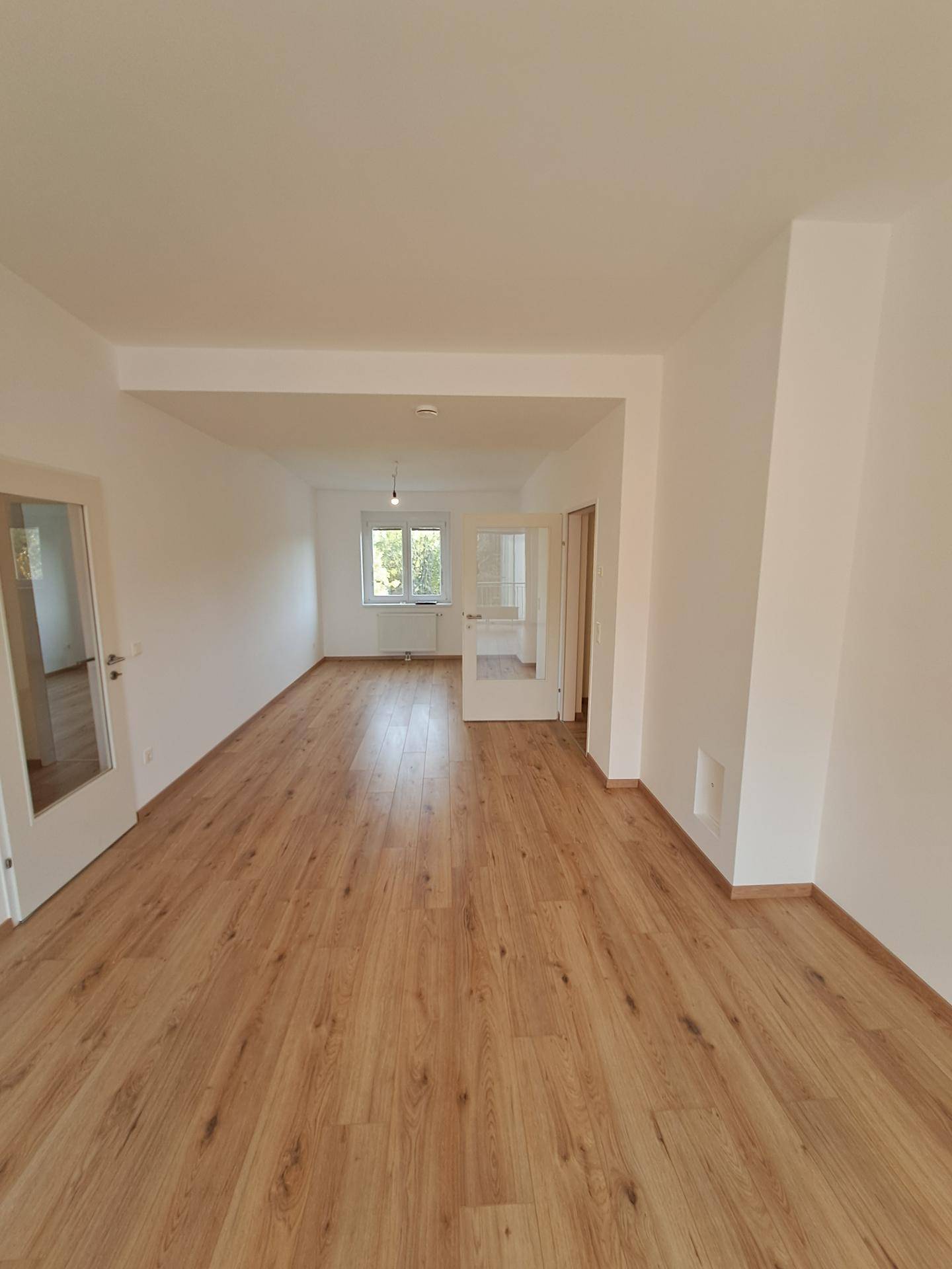 Geräumiges Wohnzimmer mit hellem Laminatboden und großen Fenstern, die viel Tageslicht hereinlassen.