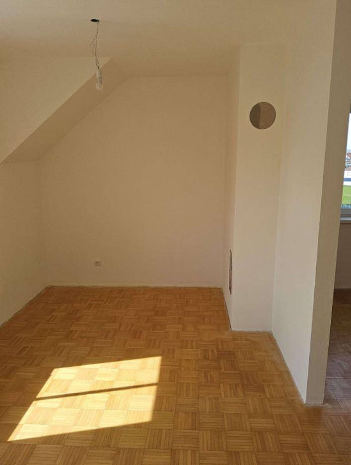 Leeres Zimmer mit Parkettboden, Dachschräge und einer runden Öffnung in der Wand.