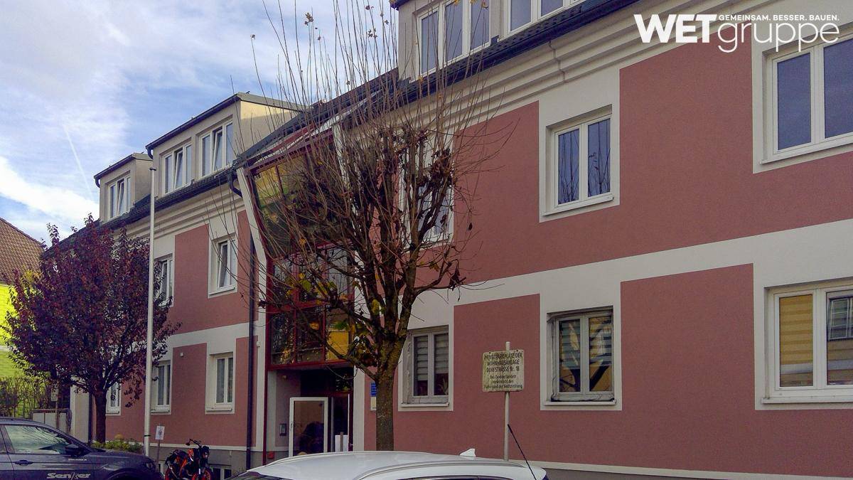 Mehrfamilienhaus mit rosa Fassade, weißen Akzenten und großen Fenstern, umgeben von Bäumen.