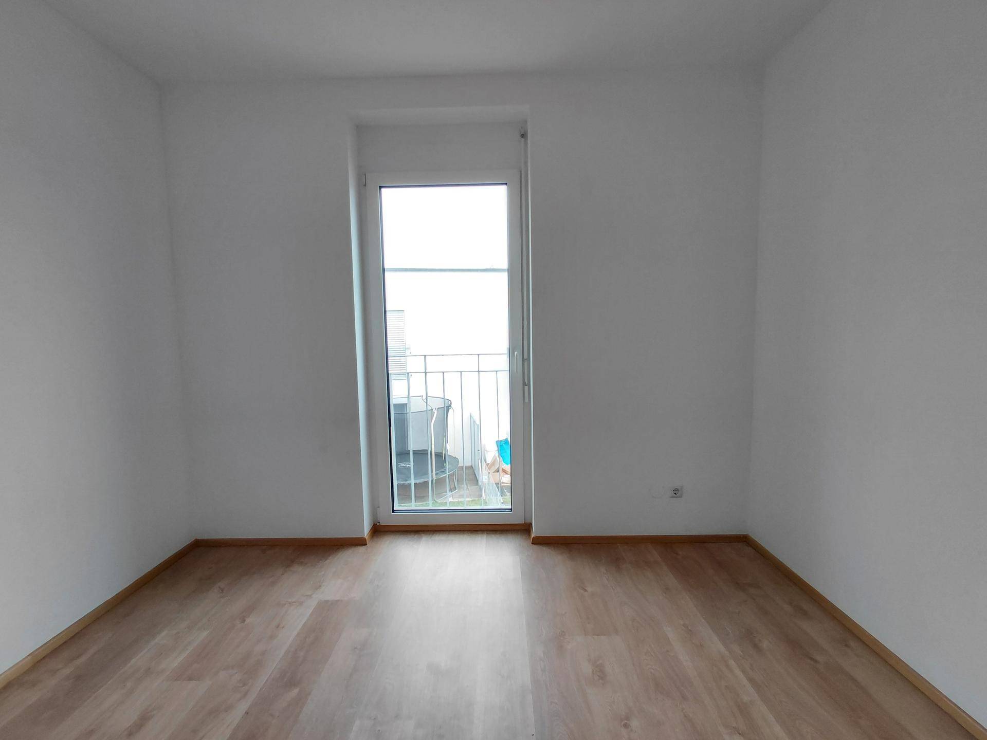 Leeres Zimmer mit Laminatboden und Zugang zu einem kleinen Balkon.