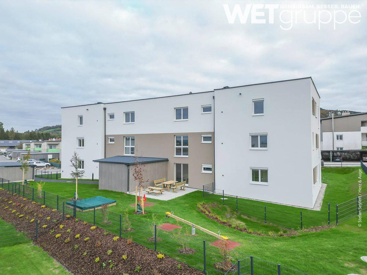 Rückseite des modernen Wohnhauses mit Gemeinschaftsgarten, Spielplatz und Sitzgelegenheiten im Freien.