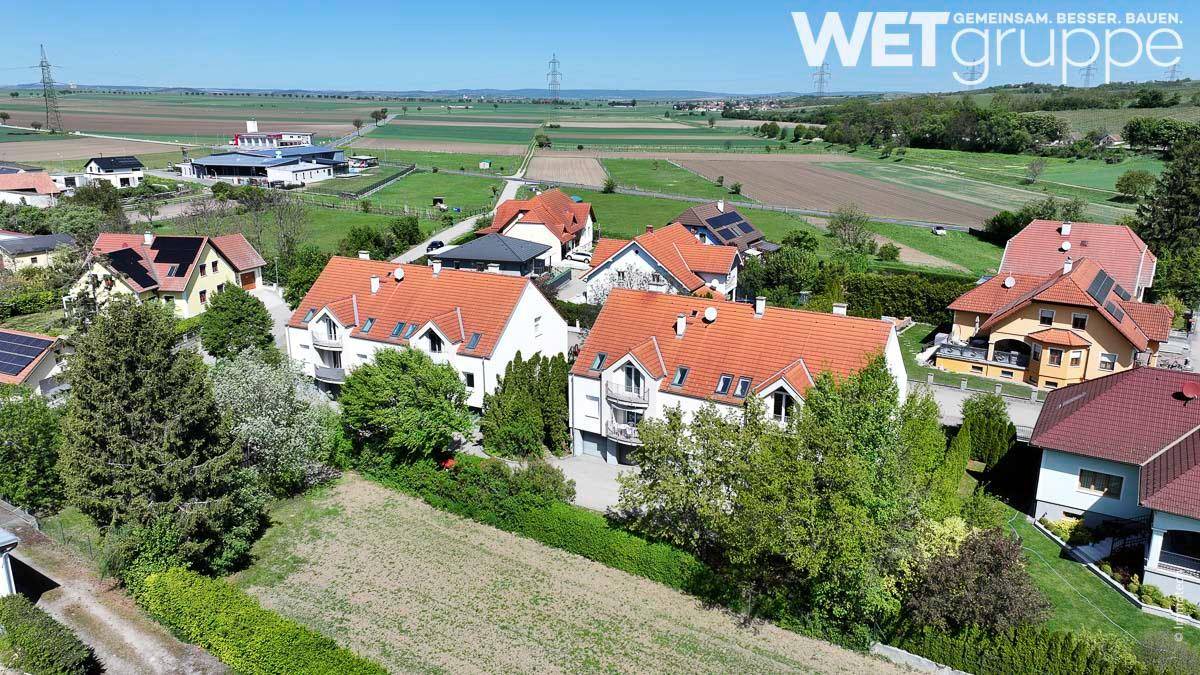 Weitläufige Luftaufnahme von Wohngebäuden in einer grünen, ländlichen Umgebung mit Feldern im Hintergrund.
