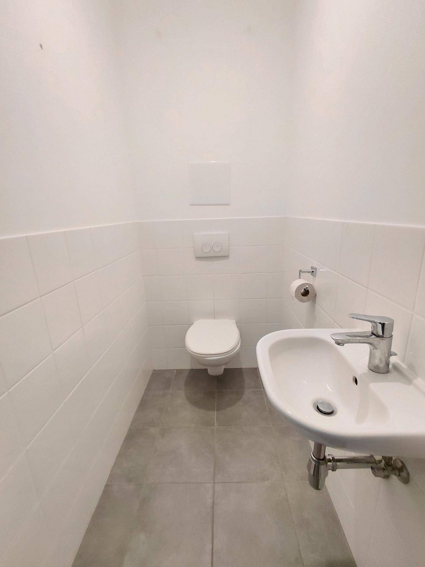 Modernes Gäste-WC mit Wand-WC, kleinem Waschbecken und grauen Bodenfliesen in sauberem Zustand.
