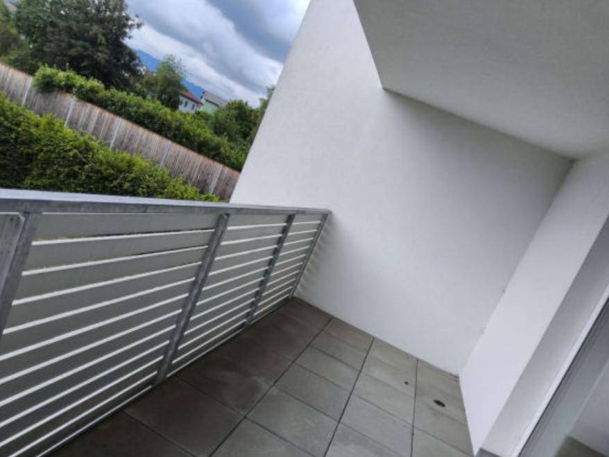Balkon mit grauen Fliesen und einer modernen Lamellenbrüstung, umgeben von grüner Bepflanzung.