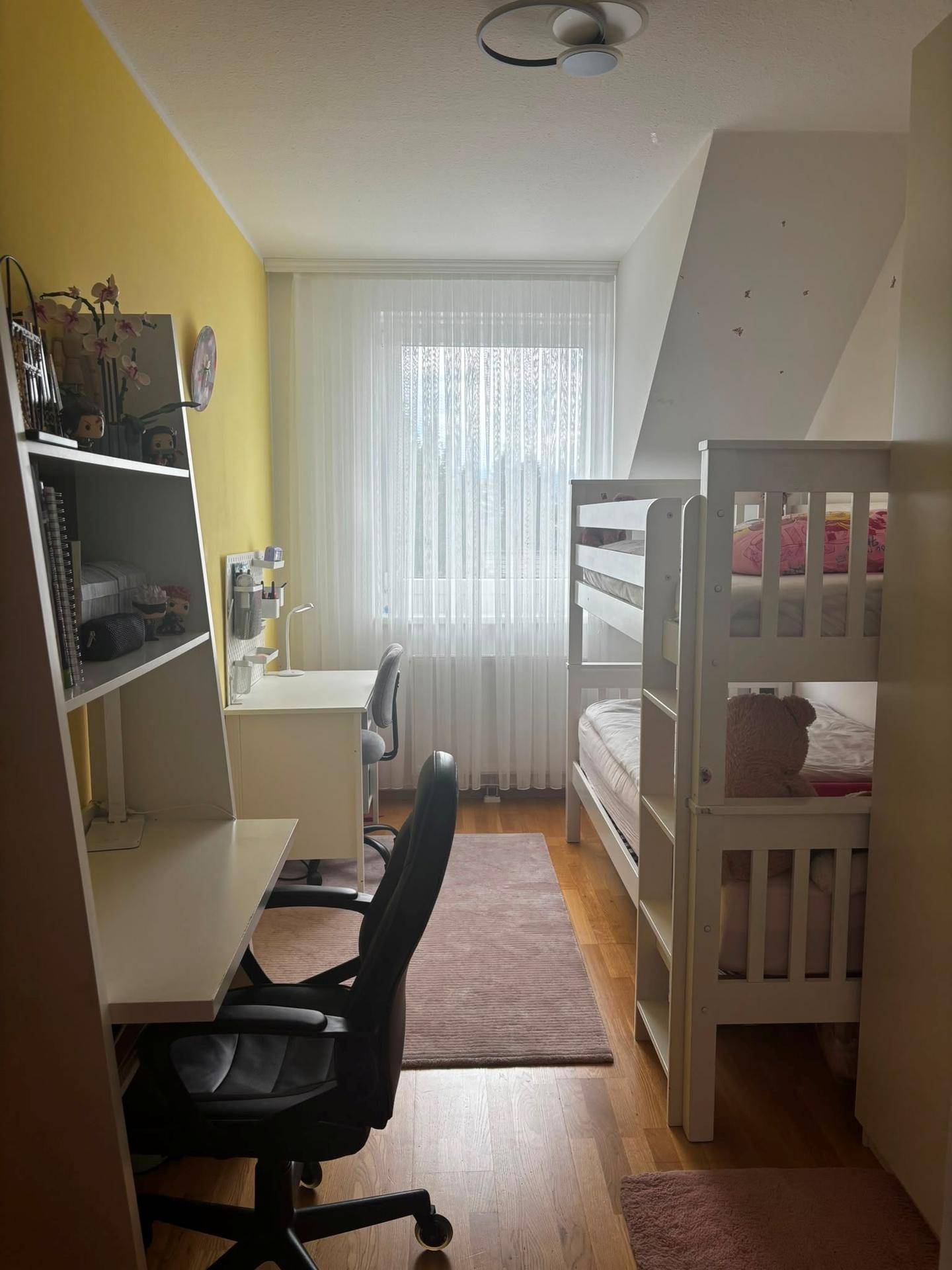 Kinderzimmer mit Etagenbett, zwei Schreibtischen und einer gelben Akzentwand, hell durch Fenster.