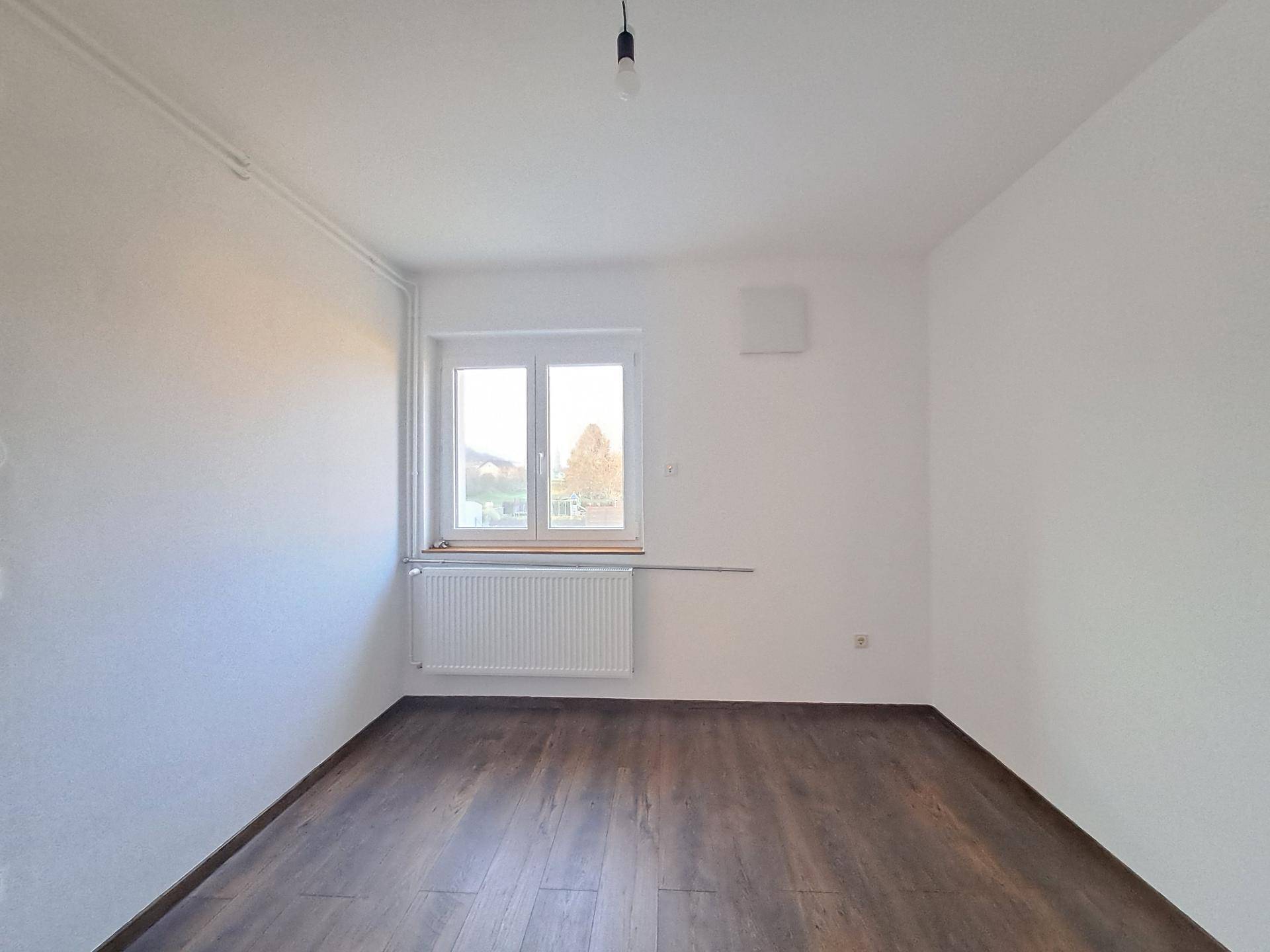 Leeres Zimmer mit dunklem Laminatboden, weißen Wänden und einem Fenster mit Heizkörper darunter.