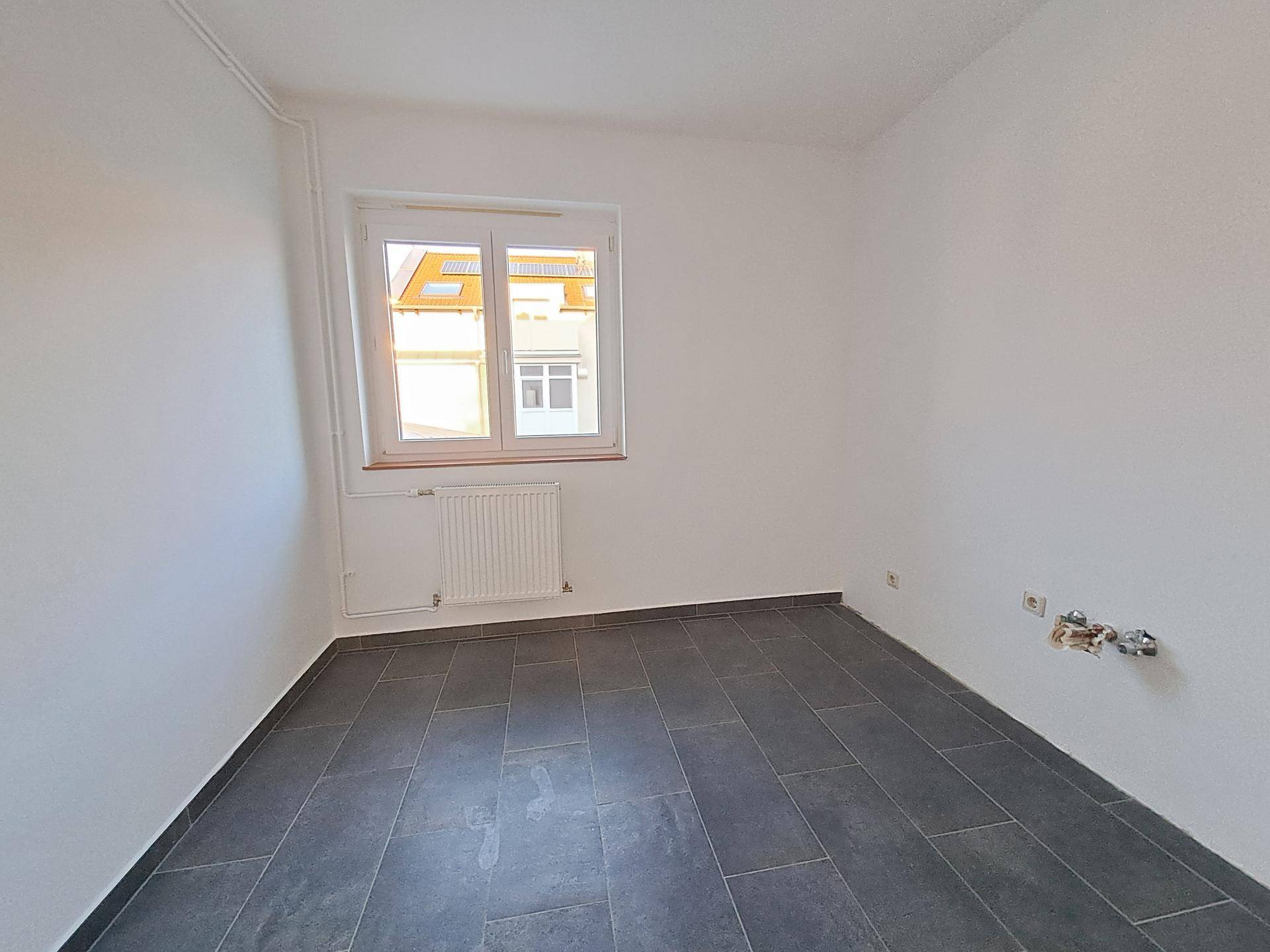 Leerer Raum mit grauem Fliesenboden, weißen Wänden und Anschlüssen für Wasserinstallationen, ideal als Waschküche.