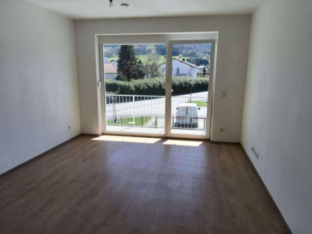 Heller Wohnraum mit Laminatboden und großem Fenster mit Zugang zu einem Balkon und Blick ins Grüne.