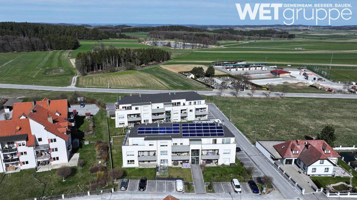 Luftbild einer modernen Wohnanlage mit Solaranlagen auf dem Dach, umgeben von ländlicher Landschaft und einem See in der Ferne.