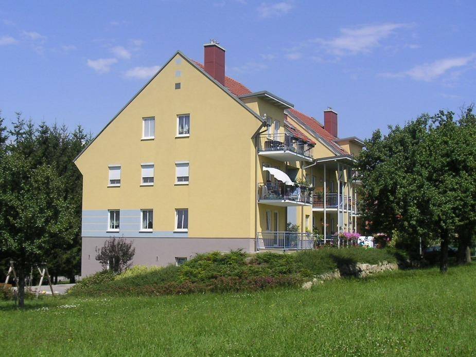 Gelbes Mehrfamilienhaus mit Balkonen, umgeben von Bäumen und einer grünen Wiese.