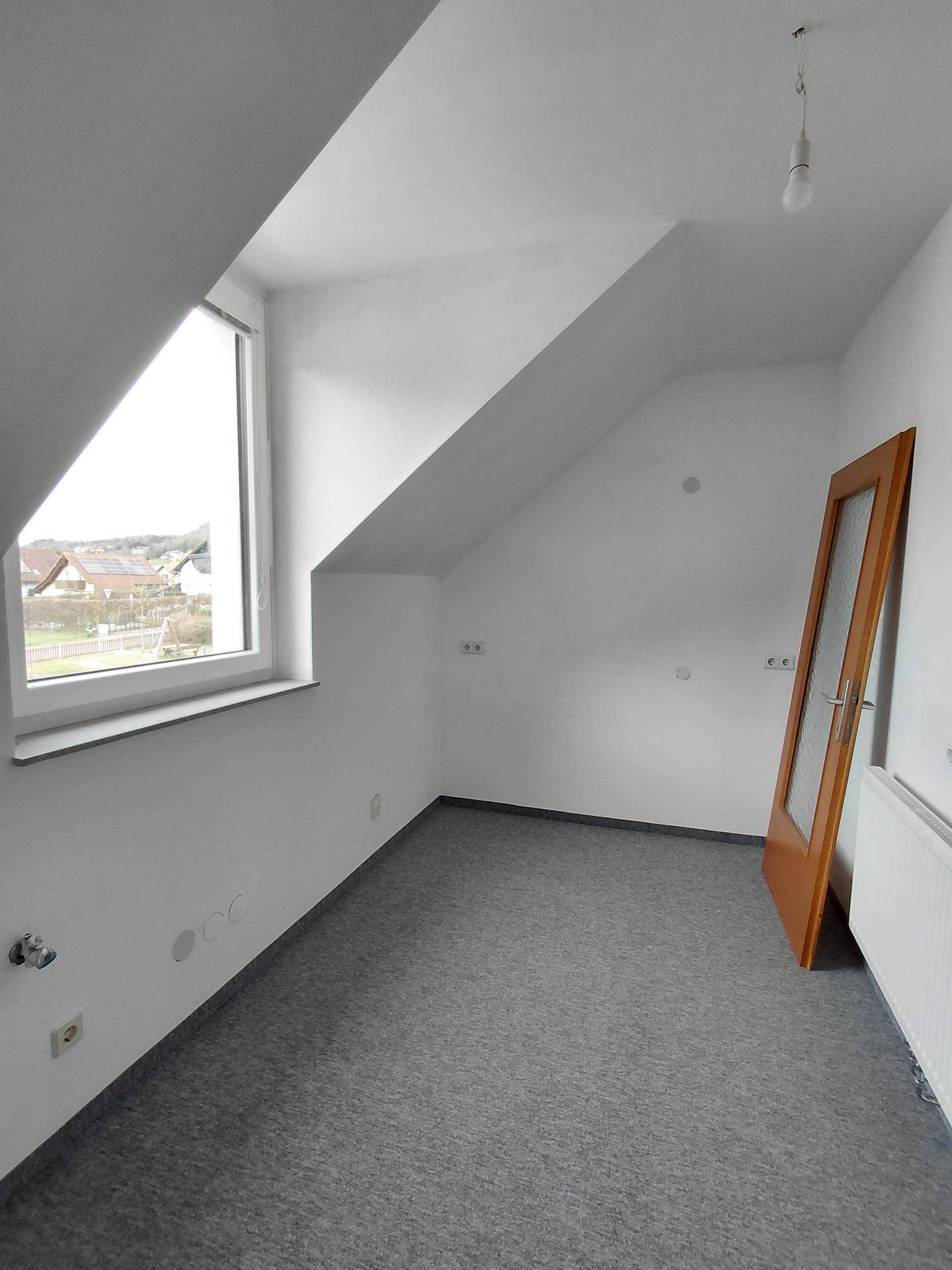 Dachzimmer mit schrägen Wänden, Fenster, Heizkörper und grauem Teppichboden, mit einer Holztür.