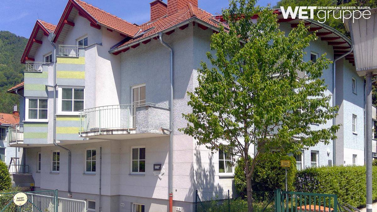 Mehrfamilienhaus mit Balkonen, rotem Ziegeldach und umgebender grüner Landschaft mit Bergen.