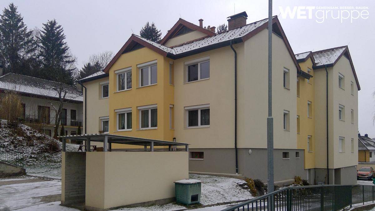Mehrfamilienhaus mit gelb-beiger Fassade und Satteldach, umgeben von Schnee und Bäumen im Winter.