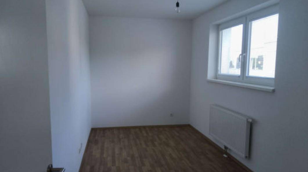 Helles, leeres Schlafzimmer mit Parkettboden und einem Fenster, das viel Tageslicht hereinlässt.