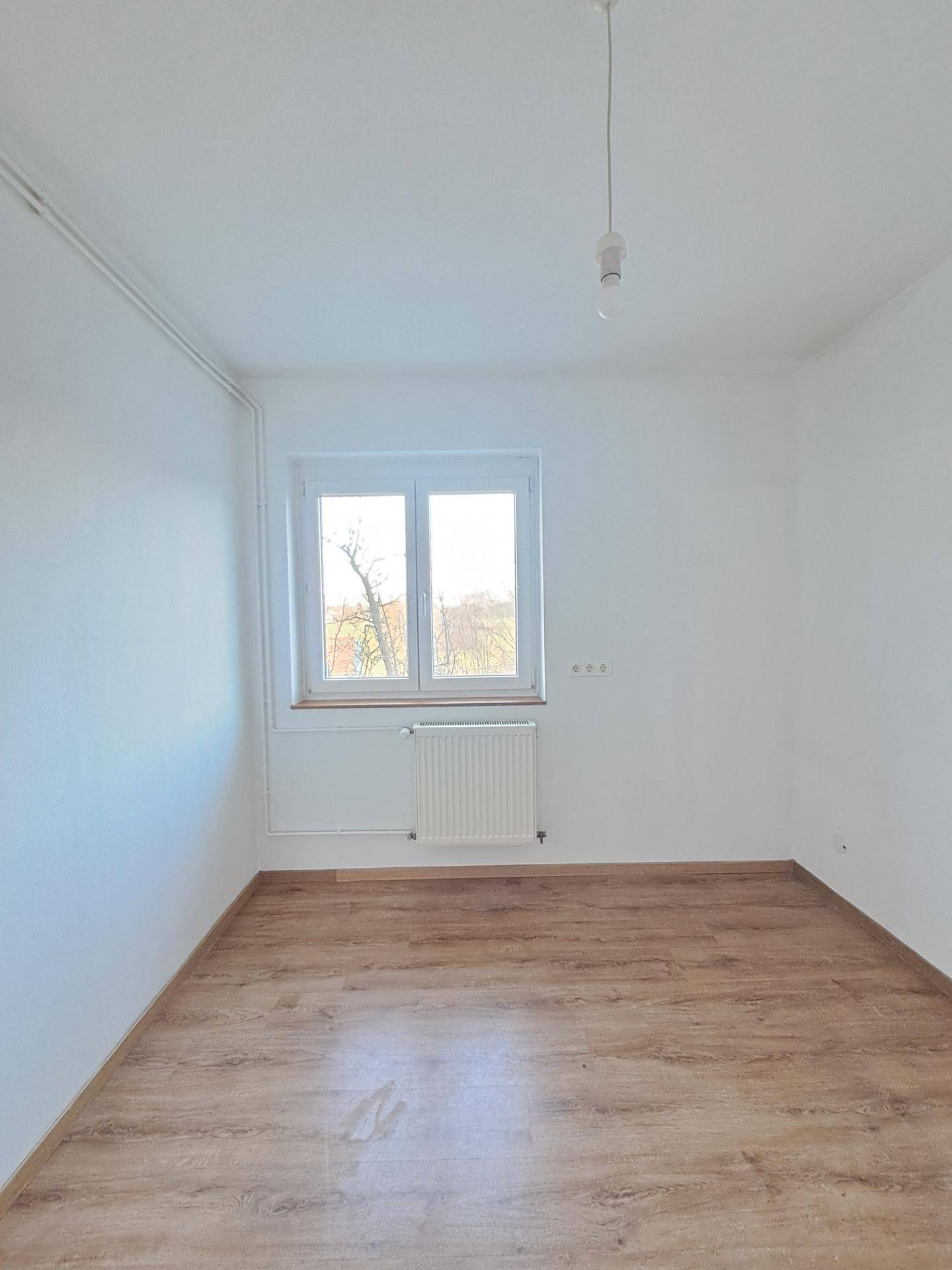 Ruhiges Zimmer mit dunklerem Laminatboden, weißen Wänden und einem Fenster für natürliches Licht.