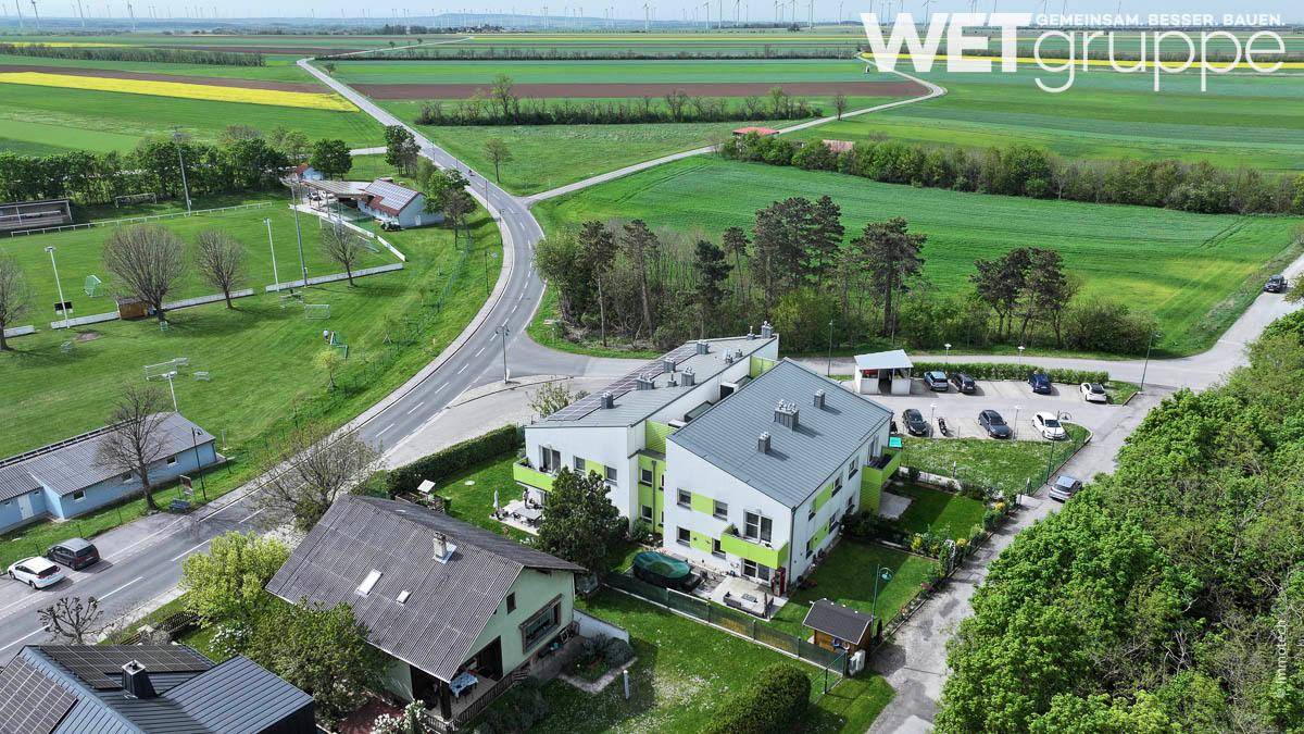 Weitläufige Luftansicht eines Wohnkomplexes in ländlicher Umgebung mit weiten Feldern und Windrädern im Hintergrund.