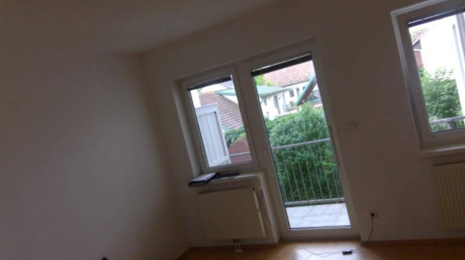 Heller Raum mit Holzboden, großen Fenstern und Zugang zu einem Balkon mit Blick ins Grüne.