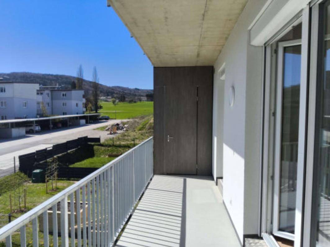 Heller Balkon mit Metallgeländer und weitem Blick über grüne Felder und benachbarte Gebäude.