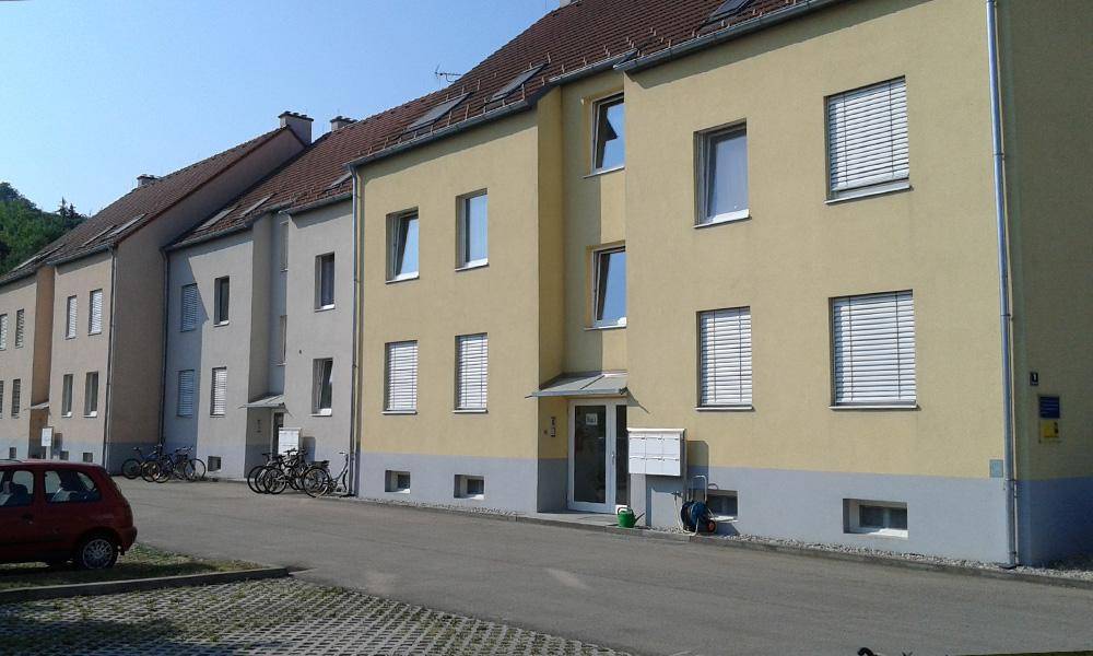 Mehrfamilienhaus mit gelber Fassade, weißen Jalousien und Parkplätzen davor.