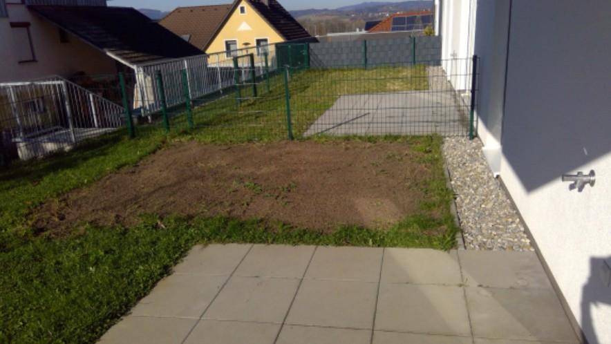 Außenbereich mit gepflasterter Terrasse, Rasenfläche und vorbereitetem Beet, umgeben von einem Zaun.