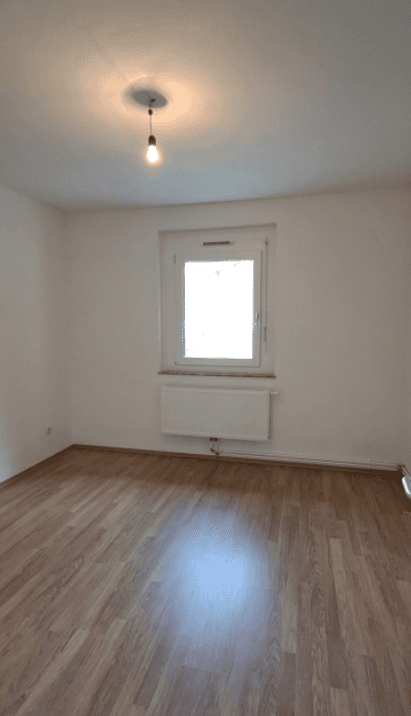 Unmöbliertes Zimmer mit Laminatboden, weiß gestrichenen Wänden und einem Fenster, das Tageslicht hereinlässt.