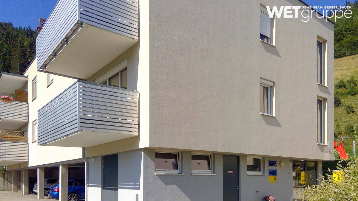 Modernes Mehrfamilienhaus mit hellgrauer Fassade, Balkonen und Tiefgaragenzugang in bergiger Umgebung.