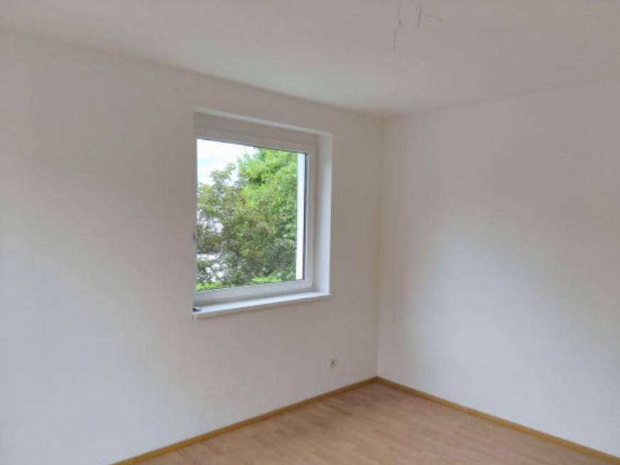 Helles Zimmer mit Holzboden, weißen Wänden und einem Fenster mit Blick ins Grüne.