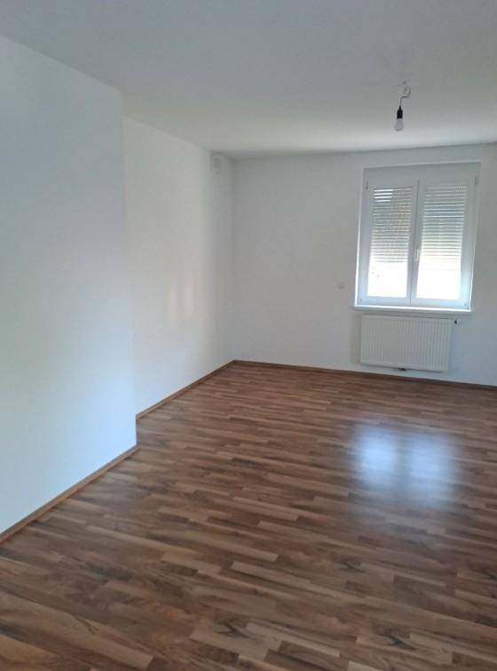 Helles, leeres Zimmer mit dunklem Laminatboden, weißen Wänden und Fenster mit Rollladen.