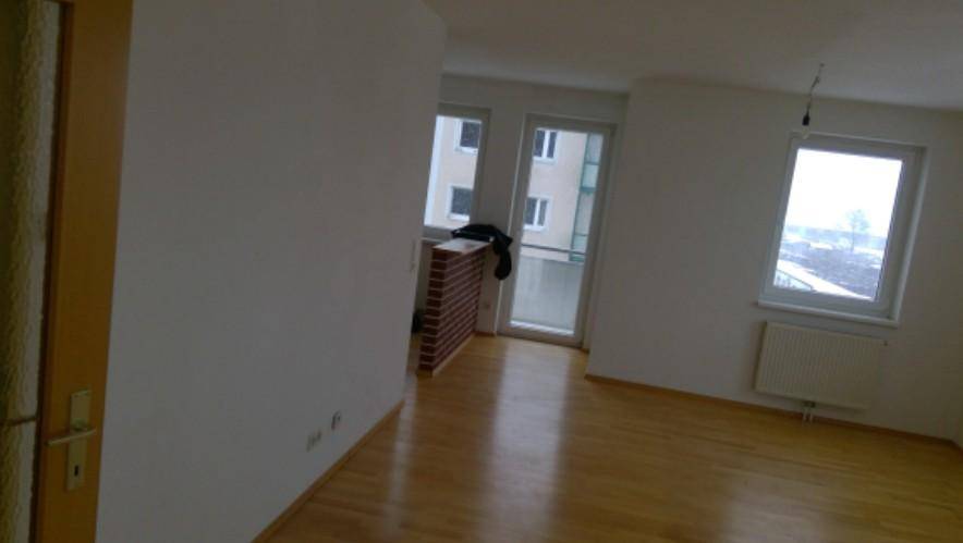Leeres Zimmer mit hellem Holzfußboden, weißen Wänden und Zugang zu einem Balkon.