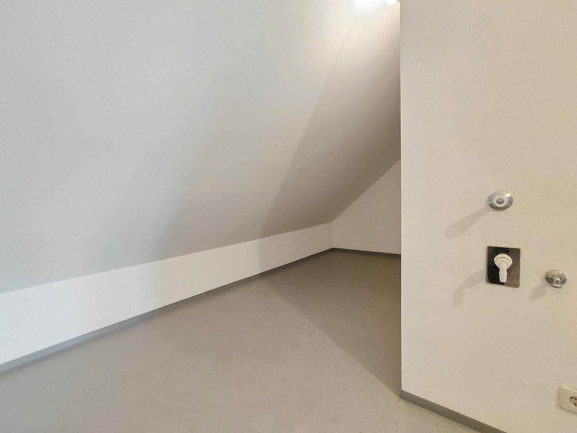 Leerer Raum im Dachgeschoss mit schrägen Wänden und grauem Bodenbelag.