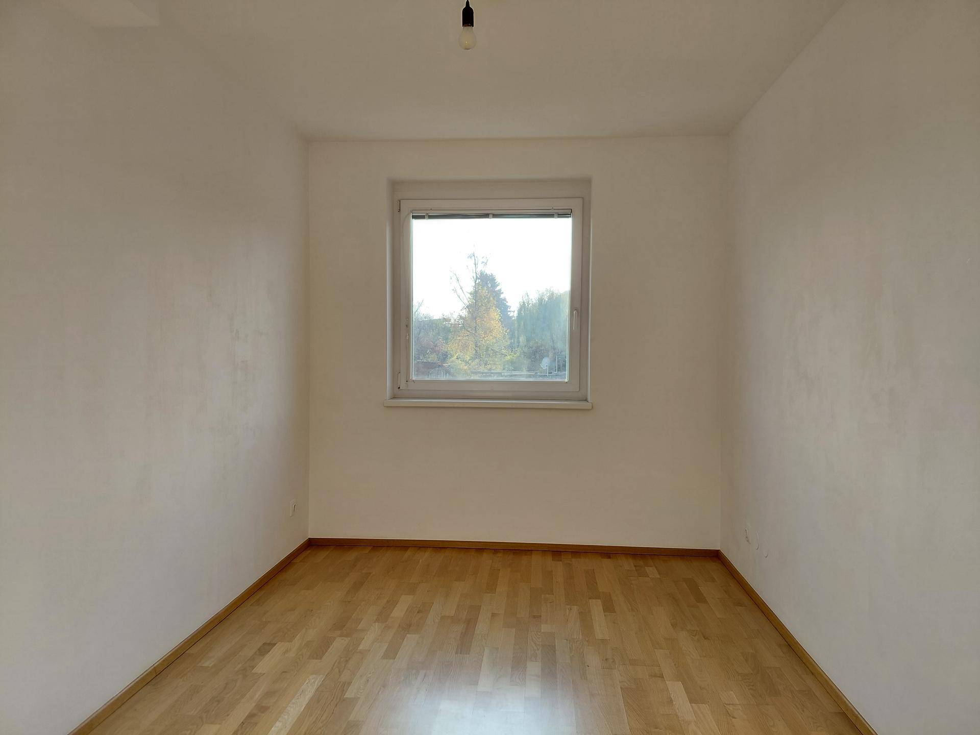 Freundliches Zimmer mit Parkettboden und Fensterblick ins Grüne, ideal für Schlaf- oder Arbeitsbereich.