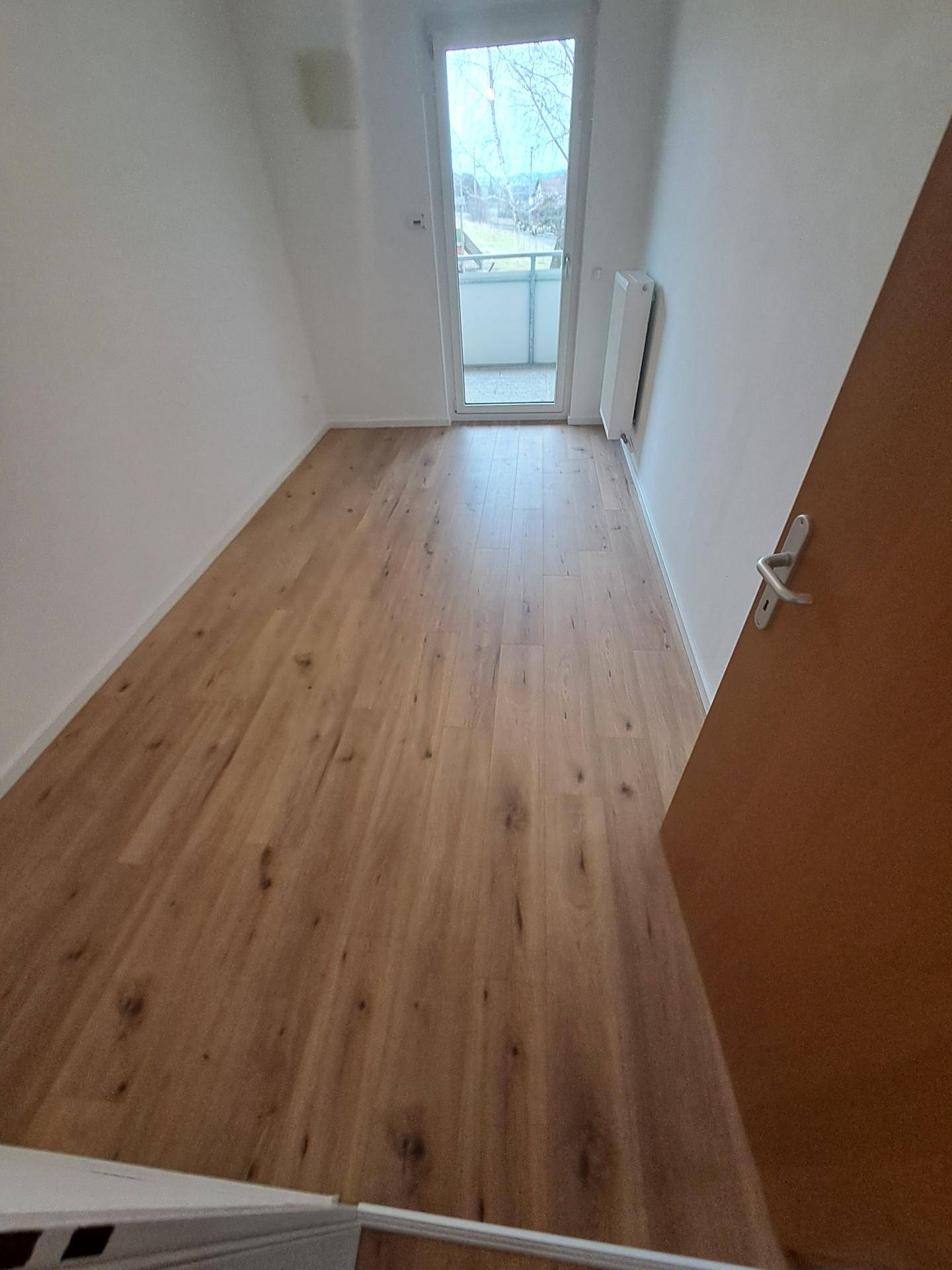 Helles Zimmer mit Holzboden und Zugang zu einem kleinen Balkon, ideal als Schlaf- oder Arbeitszimmer.