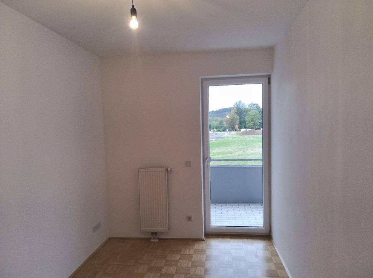 Heller Raum mit Parkettboden, Heizkörper und Zugang zu einem Balkon mit Blick ins Grüne.