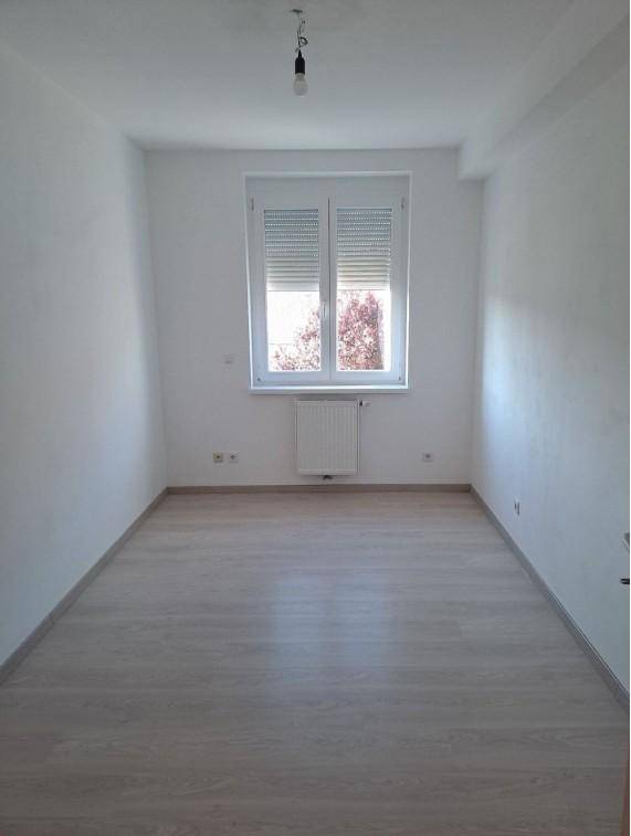 Helles, leeres Zimmer mit Laminatboden, weißen Wänden und Fenster mit Rollladen.