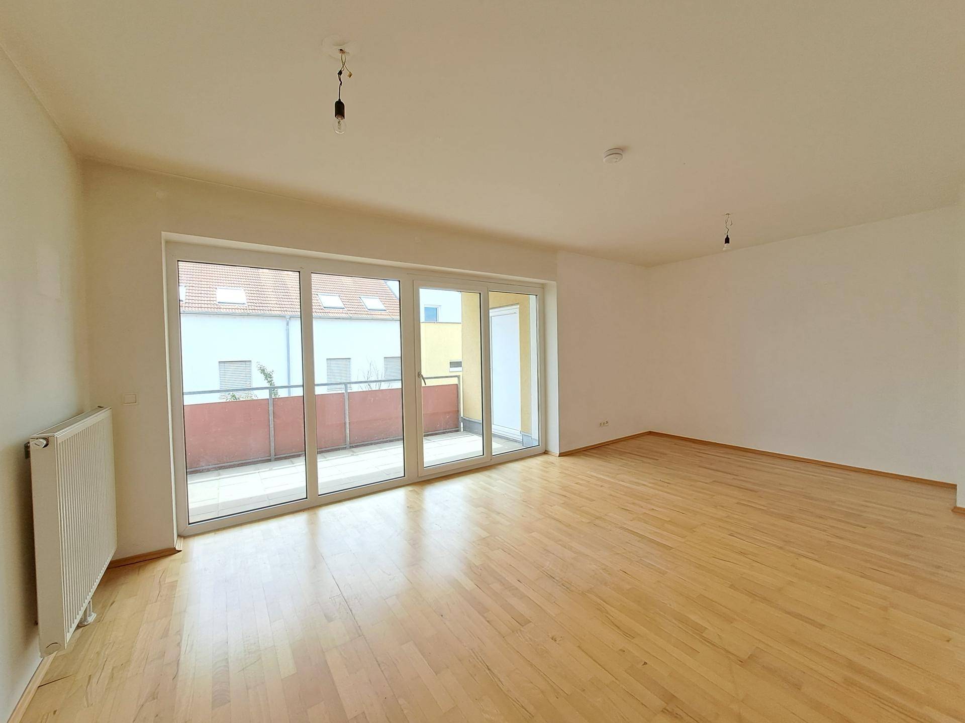 Geräumiges Wohnzimmer mit Holzboden und großen Fenstern, die Zugang zum Balkon bieten.