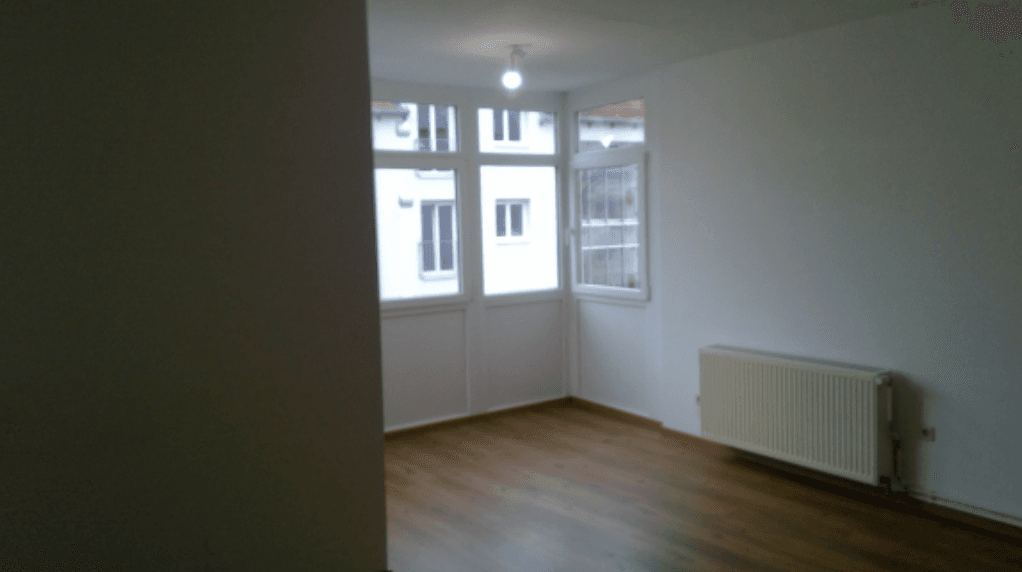 Leeres Wohnzimmer mit Holzboden, großen Fenstern und Heizkörper an der Wand.