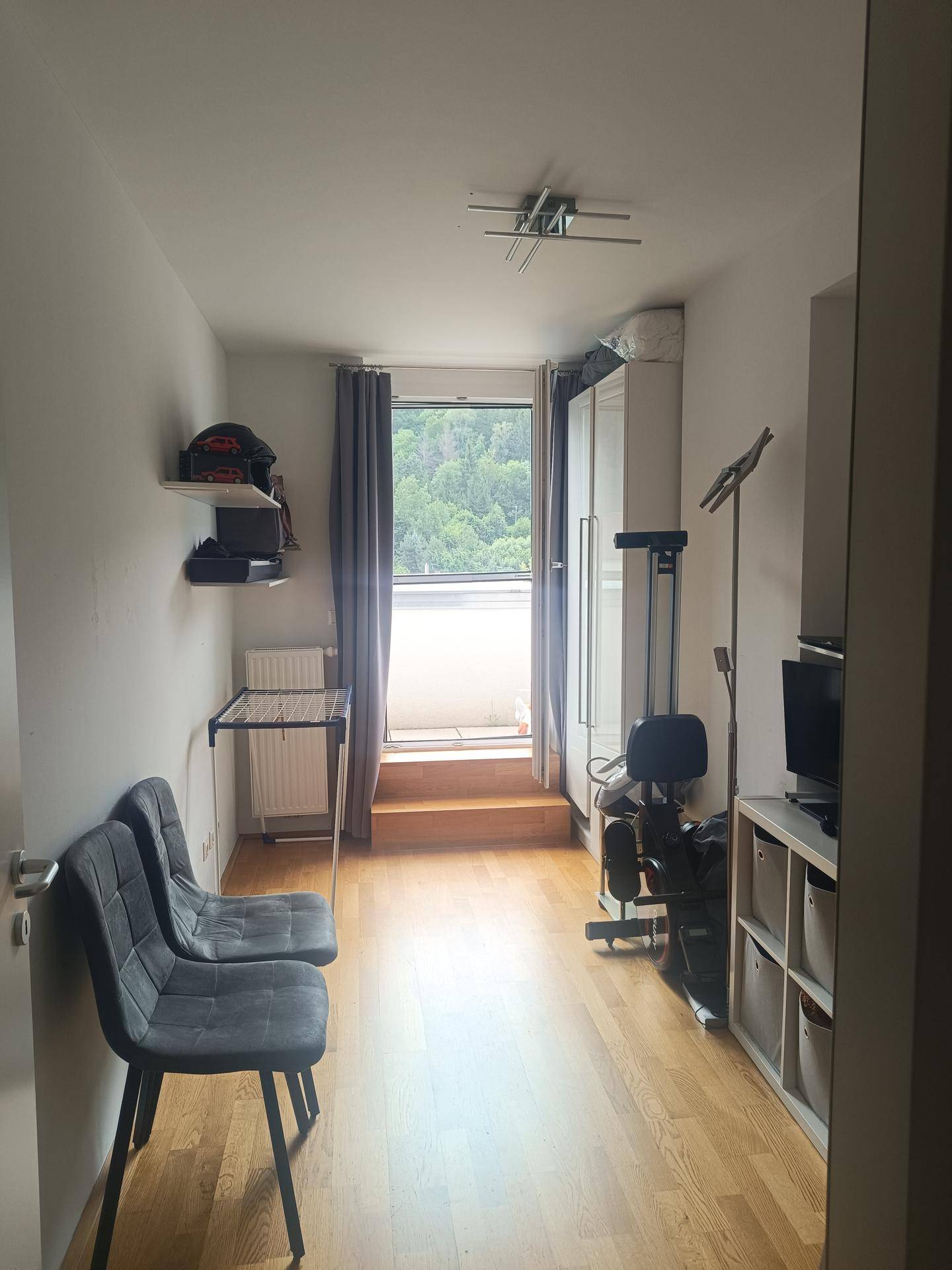 Schmaler Raum mit Holzboden und direktem Zugang zu einem Balkon mit Blick ins Grüne.