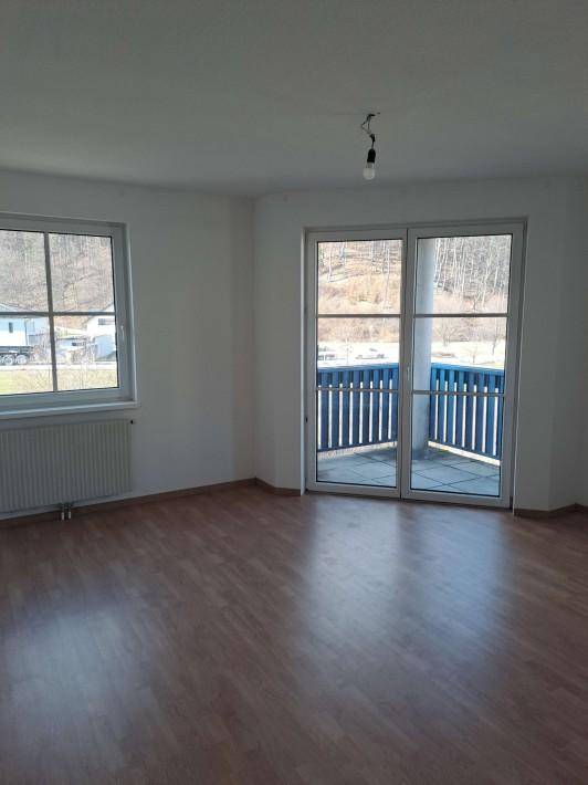 Helles Zimmer mit Holzboden, großem Fenster und Zugang zu einem Balkon mit blauem Geländer.