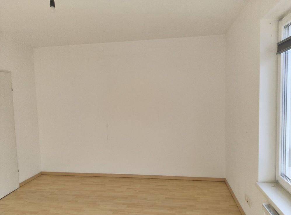 Leerer Raum mit hellen Wänden, Holzboden und einem Fenster, das Tageslicht hereinlässt.