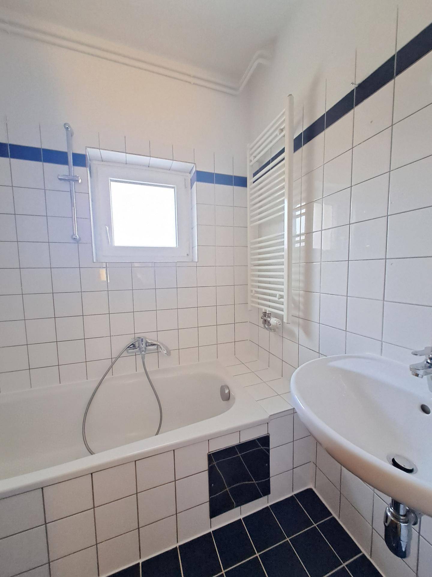 Helles Badezimmer mit Badewanne, Waschbecken, Fenster und Handtuchheizkörper in weiß-blauer Farbgebung.